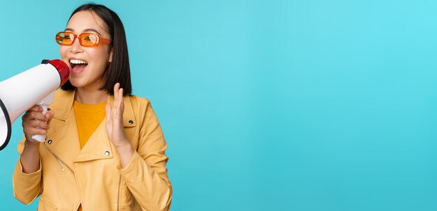 Stilvolles asiatisches Mädchen, das Ankündigung im Megaphon macht, das mit Freisprecheinrichtung schreit und lächelnde einladende Leute rekrutiert, die über blauem Hintergrund stehen