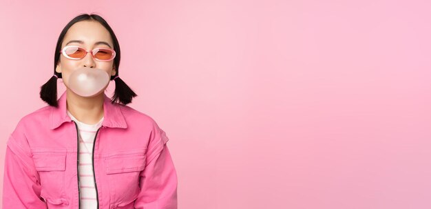 Stilvolles asiatisches Mädchen, das Bubblegum-Kaugummi bläst und eine Sonnenbrille trägt, die vor rosa Hintergrund posiert