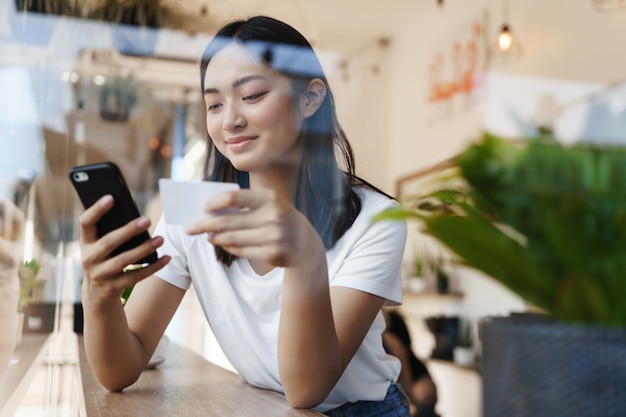 Stilvolles asiatisches Mädchen, das in einem Café nahe dem Fenster sitzt und für Online-Einkauf mit einer Kreditkarte bezahlt.