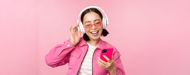 Stilvolles asiatisches Mädchen, das mit dem Smartphone tanzt und Musik in Kopfhörern auf der Handy-App hört, lächelt und lacht und posiert vor rosa Hintergrund