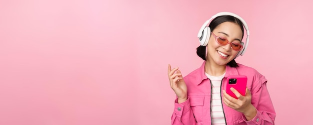 Stilvolles asiatisches Mädchen, das mit dem Smartphone tanzt und Musik in Kopfhörern auf der Handy-App lächelt