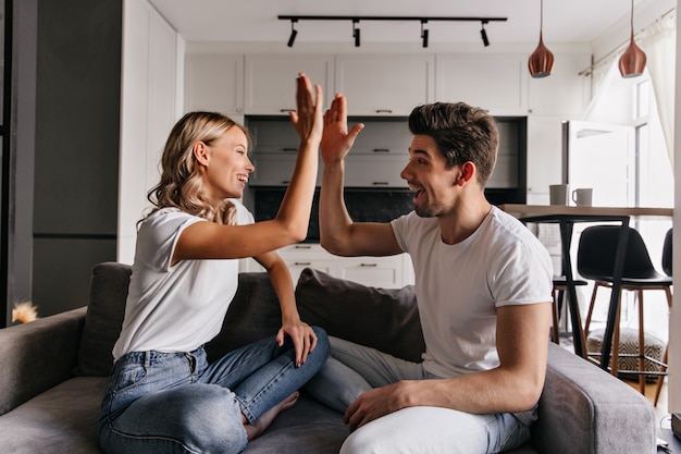 Stilvolles blondes Mädchen, das mit Freund im Wohnzimmer spielt. Innenporträt von scherzenden jungen Leuten.