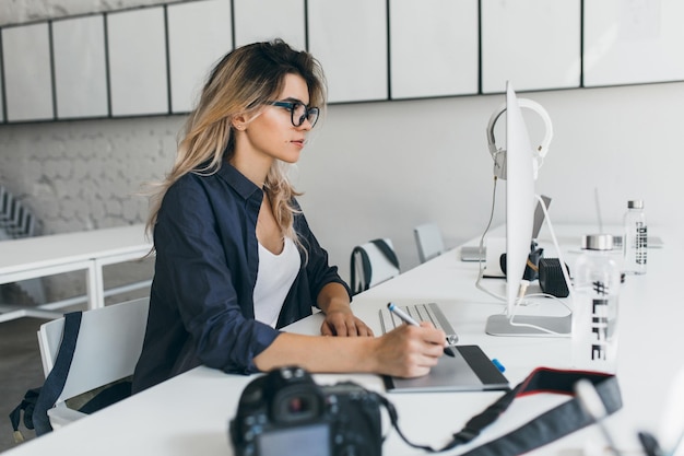 Stilvolles blondes Mädchen, das nach dem Fotoshooting Tablet benutzt und mit Computer und Kamera am Tisch sitzt. Charmante Studentin in Brille und schwarzem Hemd, die an einem Projekt arbeitet.