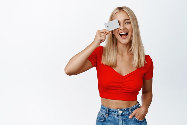 Stilvolles blondes Mädchen zeigt Kreditkarte nahe Gesicht und lachendes Konzept des Einkaufens und des kontaktlosen Bezahlens, das über weißem Hintergrund steht