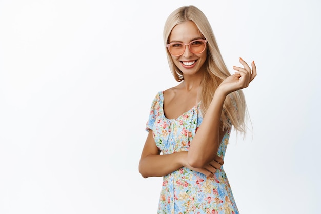 Stilvolles blondes Sommermädchen mit Sonnenbrille und Kleid lächelnd und kokett in die Kamera schauend, die über weißem Hintergrund steht