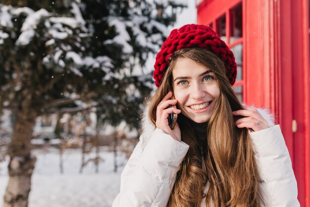 Stilvolles britisches Porträt der erstaunlichen jungen Frau mit dem langen brünetten Haar im roten Hut, der am Telefon auf der Straße voll mit Schnee spricht. Genießen Sie die kalte Winterzeit und die fröhliche Stimmung. Platz für Text.