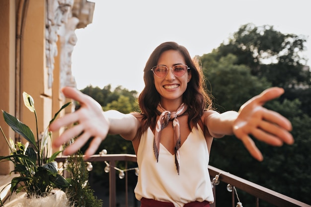 Stilvolles brünettes Mädchen, das lächelt und für Kamera greift. Moderne Frau in der rosa Sonnenbrille in der weißen Bluse, die auf Terrasse aufwirft.