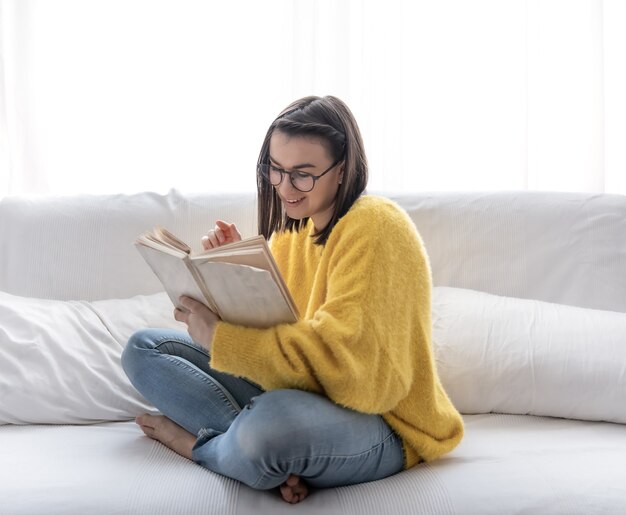 Stilvolles brünettes Mädchen in einem gelben Pullover und in den Gläsern liest ein Buch zu Hause auf der Couch. Das Konzept der Selbstentwicklung und Entspannung.