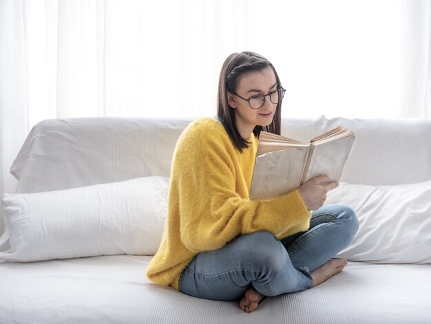 Stilvolles brünettes Mädchen in einem gelben Pullover und in den Gläsern liest ein Buch zu Hause auf der Couch.