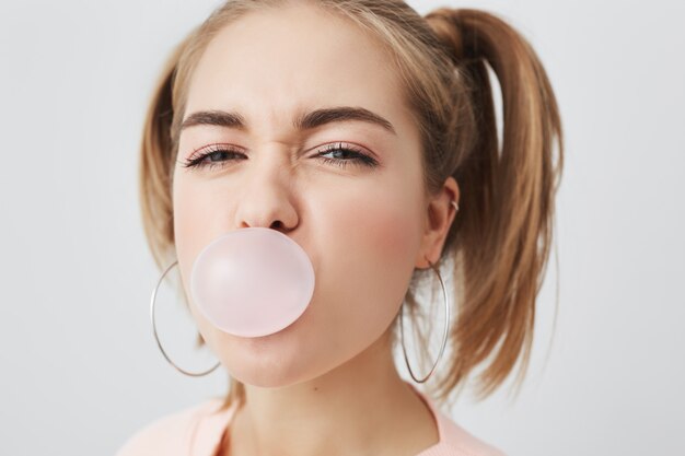 Stilvolles charmantes schönes Teenager-Mädchen mit zwei Pferdeschwänzen, die ihre Augen zusammenziehen, mit rosa Kaugummi im Mund, Freude drinnen habend. Schöne positive junge Frau in Rosa, die Spaß drinnen hat