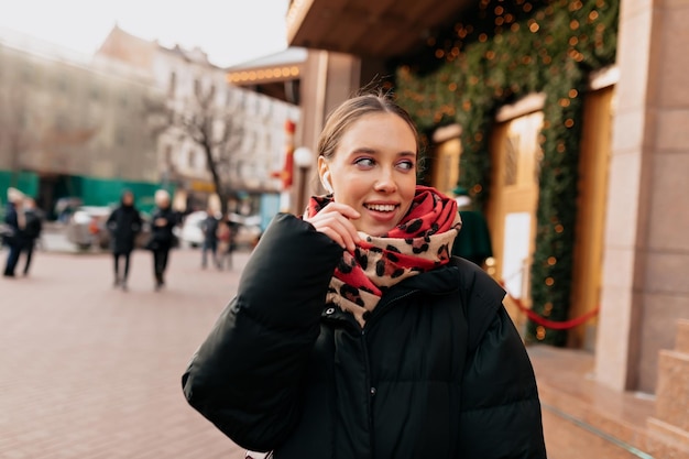 Stilvolles europäisches dunkelhaariges Mädchen mit hellem Make-up in dunkler Jacke hört Musik im Kopfhörer, schaut zur Seite und geht die Straße im Stadtzentrum hinunter