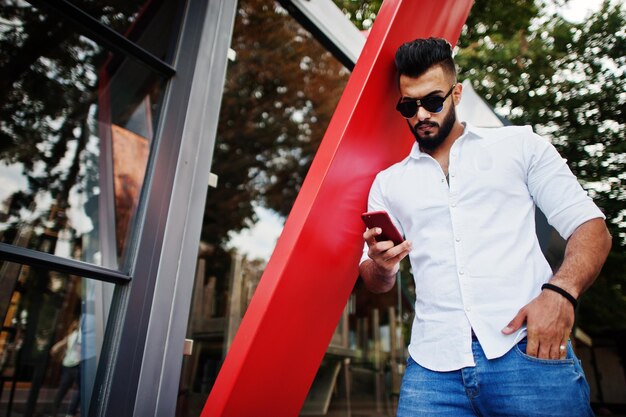 Stilvolles großes arabisches Mannmodell in weißer Hemdjeans und Sonnenbrille posierte auf der Straße der Stadt Bart attraktiver arabischer Kerl, der auf rotes Handy schaut