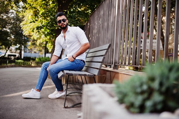 Stilvolles großes arabisches Mannmodell in weißer Hemdjeans und Sonnenbrille posierte auf der Straße der Stadt Bart attraktiver arabischer Typ, der auf einer Bank sitzt