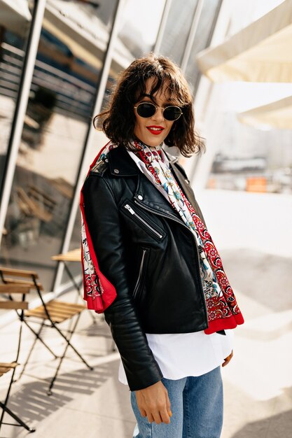Stilvolles Hipster-Mädchen, das Lederjacke mit kurzer Frisur und rotem Lippenstift trägt, die in der Stadt im Sonnenlicht gehen