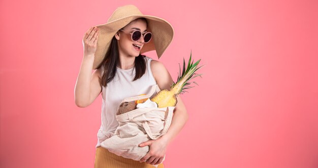 Stilvolles junges Mädchen mit großem Hut und Sonnenbrille lächelt und hält eine Öko-Tasche mit exotischen Früchten