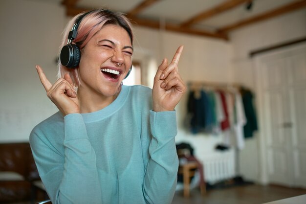 Stilvolles junges Mädchen mit rosa Haaren und Piercing im Gesicht, das Spaß drinnen hat, allein zu Hause ist, Musik in drahtlosen Kopfhörern hört, Augen schließt, Tanzbewegungen macht, mitsingt