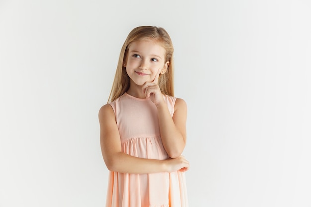Stilvolles kleines lächelndes Mädchen, das im Kleid lokalisiert auf weißem Studiohintergrund aufwirft. Kaukasisches blondes weibliches Modell. Menschliche Gefühle, Gesichtsausdruck, Kindheit. Denken oder träumen,