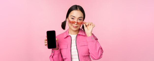 Stilvolles koreanisches Mädchen, junge Frau mit Sonnenbrille, die Smartphone-Bildschirm-Handy-App-Schnittstelle oder Website zeigt, die über rosa Hintergrund steht
