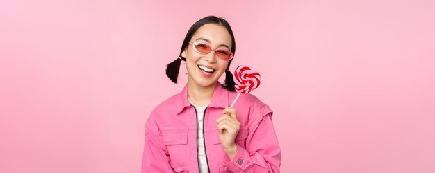 Stilvolles koreanisches Mädchen leckt Lolipop, isst Süßigkeiten und lächelt in Sonnenbrillen vor rosa Hintergrund