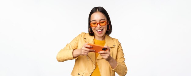 Stilvolles koreanisches Mädchen mit Sonnenbrille, das ein mobiles Videospiel spielt, lacht und lächelt, während es das Smartphone benutzt, das über weißem Hintergrund steht