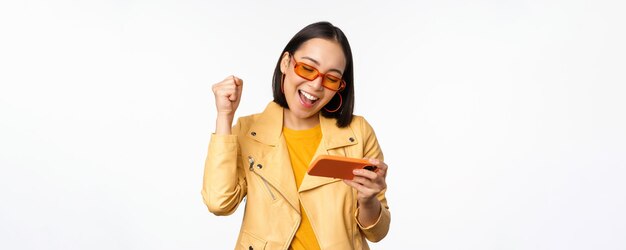 Stilvolles koreanisches Mädchen mit Sonnenbrille, das ein mobiles Videospiel spielt, lacht und lächelt, während es das Smartphone benutzt, das über weißem Hintergrund steht