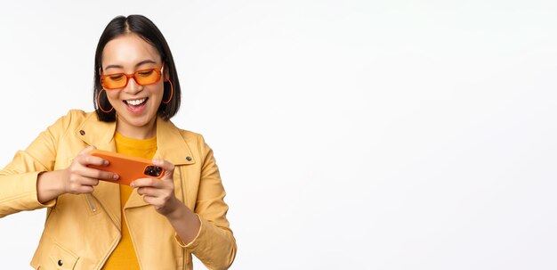 Stilvolles koreanisches Mädchen mit Sonnenbrille, das ein mobiles Videospiel spielt, lacht und lächelt, während es Smartph verwendet