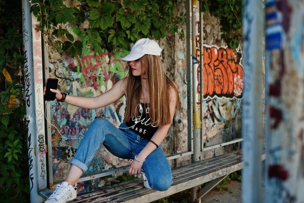 Stilvolles, lässiges Hipster-Mädchen in Mütze und Jeans, das Musik von Kopfhörern des Mobiltelefons an einer großen Graffitiwand hört