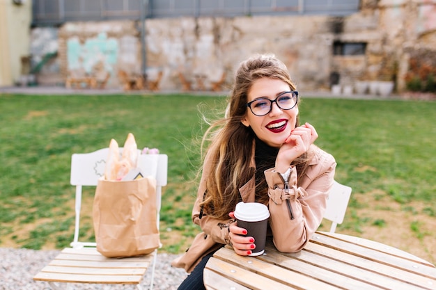 Stilvolles langhaariges Mädchen, das braunen Mantel und Gläser trägt, trinkt Latte im Café nach dem Einkaufen mit Taschen auf Stuhl hinter. Kaffeepause im Außenrestaurant auf dem unscharfen Hintergrund.