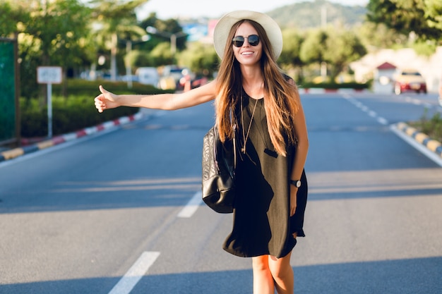 Stilvolles Mädchen, das auf der Straße per Anhalter fährt und kurzes schwarzes Kleid, Strohhut, schwarze Brille und schwarzen Rucksack trägt. Sie lächelt in den warmen Strahlen der untergehenden Sonne