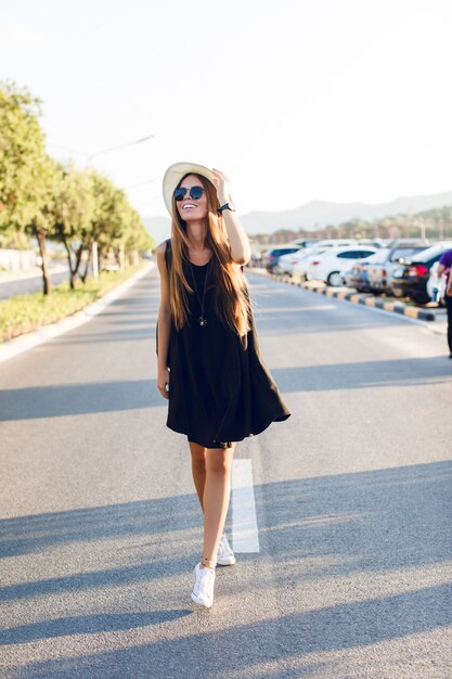 Stilvolles Mädchen, das in der Nähe der Straße steht und kurzes schwarzes Kleid, Strohhut, schwarze Brille, weiße Turnschuhe und schwarzen Rucksack trägt. Sie lächelt in den warmen Strahlen der untergehenden Sonne