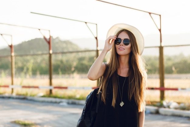 Stilvolles Mädchen, das nahe Straße steht und kurzes schwarzes Kleid, Strohhut, schwarze Brille und schwarzen Rucksack trägt. Sie lächelt in den warmen Strahlen der untergehenden Sonne. Sie berührt ihre Sonnenbrille mit der Hand