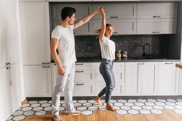 Stilvolles Mädchen in den Jeans, die mit Ehemann am Morgen tanzen. Innenporträt der entspannten jungen Leute, die Spaß in der Küche haben.