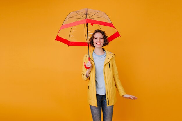 Stilvolles Mädchen in den Jeans und im Regenmantel, die unter niedlichem Regenschirm stehen. Innenporträt der romantischen jungen Frau mit der lockigen Frisur, die Sonnenschirm auf orange Wand hält.