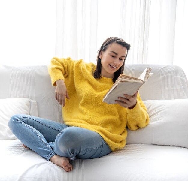 Stilvolles Mädchen in einem gelben Pullover ruht zu Hause auf dem Sofa mit einem Buch.