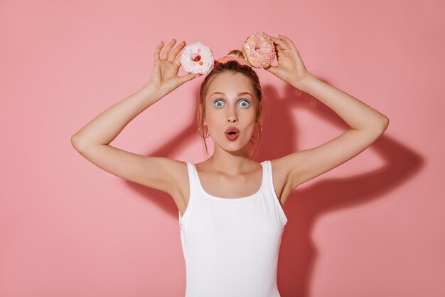 Stilvolles Mädchen mit modernem Make-up und goldenen Ohrringen in hellem Badeanzug, das in die Kamera schaut und mit Donuts auf rosafarbenem Hintergrund posiert