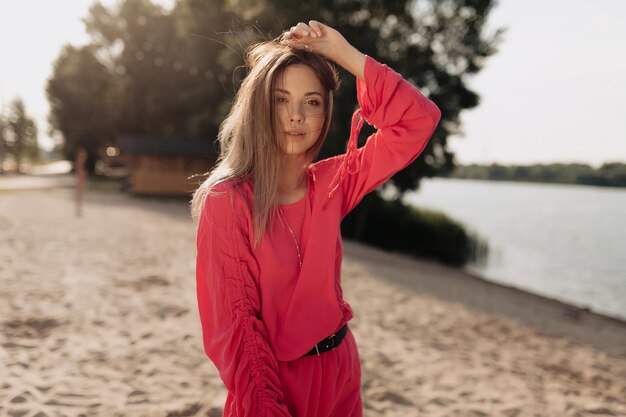 Stilvolles Model mit fliegendem, dunkelhaarigem, rosafarbenem Sommerkleid blickt in die Kamera und berührt ihr Haar auf dem Hintergrund des Sandstrandes bei Sonnenschein