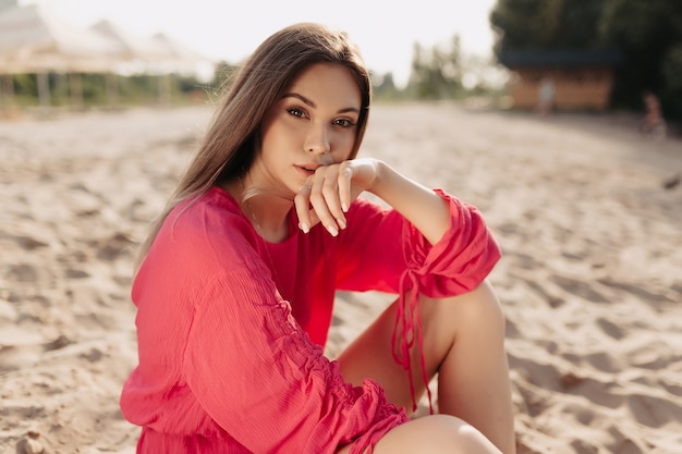 Stilvolles modernes weibliches Modell im rosa Sommerkleid, das auf dem Strand im Sonnenlicht aufwirft