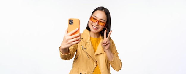 Stilvolles schönes asiatisches Mädchen mit Sonnenbrille, das Selfie auf dem Smartphone macht und für ein Foto posiert, das den weißen Studiohintergrund des Mobiltelefons hält