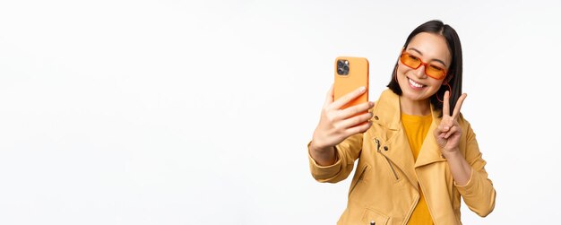 Stilvolles schönes asiatisches Mädchen mit Sonnenbrille, das Selfie auf dem Smartphone macht und für ein Foto posiert, das den weißen Studiohintergrund des Mobiltelefons hält