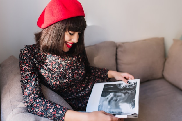 Stilvolles spektakuläres mädchen in roter baskenmütze schaut interessiert auf das fotobuch und lehnt ihre ellbogen auf ein graues sofa. porträt der bezaubernden jungen französischen frau im weinlesemagazin der weinlesekleidung in der freizeit