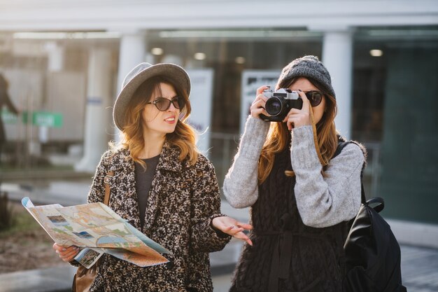 Stilvolles Stadtporträt von zwei modischen Frauen, die im modernen Stadtzentrum Europas gehen. Modische Freunde, die mit Rucksack, Karte, Kamera reisen, Fotos machen, Touristen, verlieren sich.