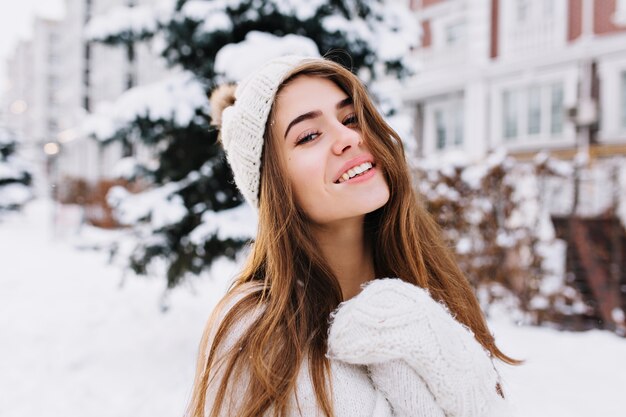 Stilvolles Winterporträt der bezaubernden jungen Frau mit dem langen brünetten Haar, in den weißen Wollhandschuhen und im Hut, die auf der Straße voll mit Schnee gehen. Fröhliche Stimmung, lächelnd.