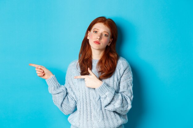 Stimmungsvolles Teenager-Mädchen mit roten Haaren, das mit den Fingern nach links auf das Logo zeigt, belästigt und gelangweilt starrt, auf blauem Hintergrund stehend.