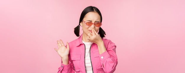 Stinkendes asiatisches Mädchen mit Sonnenbrille und stylischem Outfit schließt ihre Nase vor Ekel, Abneigung gegen schlechten Geruch, der über rosafarbenem Hintergrund steht