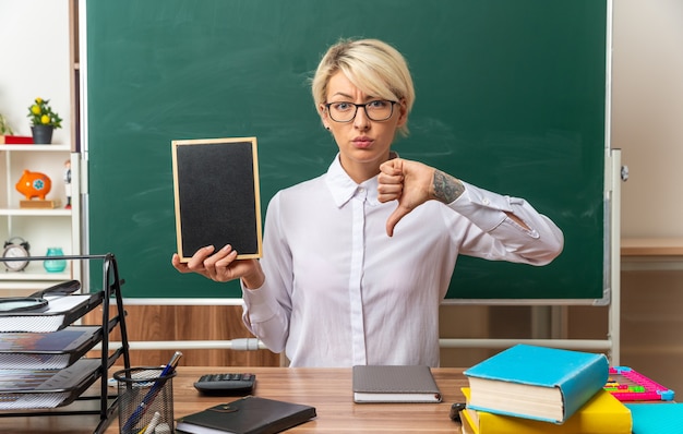 Stirnrunzelnd junge blonde Lehrerin mit Brille am Schreibtisch sitzend mit Schulwerkzeugen im Klassenzimmer mit Mini-Tafel und Daumen nach unten