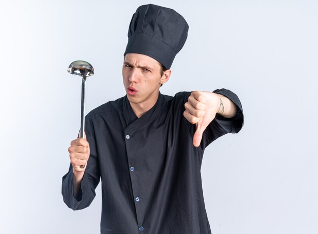 Stirnrunzelnd junger blonder männlicher Koch in Kochuniform und Mütze mit Schöpfkelle mit Blick in die Kamera mit Daumen nach unten isoliert auf weißer Wand