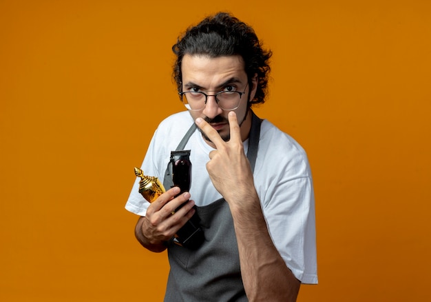 Stirnrunzelnd junger kaukasischer männlicher barbier mit brille und welligem haarband in uniform, der gewinnerbecher und haarschneidemaschinen hält, ich beobachte deine geste