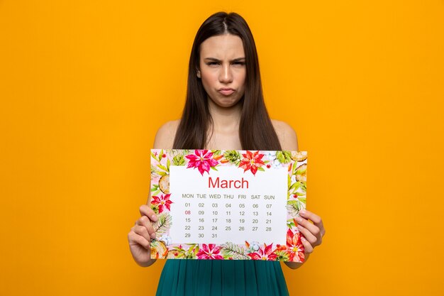 Stirnrunzelnd schönes junges Mädchen am Tag der glücklichen Frau mit Kalender isoliert auf oranger Wand