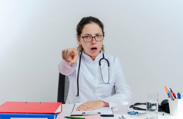 Stirnrunzelnde junge Ärztin, die medizinische Robe und Stethoskop und Brille trägt, die am Schreibtisch mit medizinischen Werkzeugen sitzen und lokalisieren