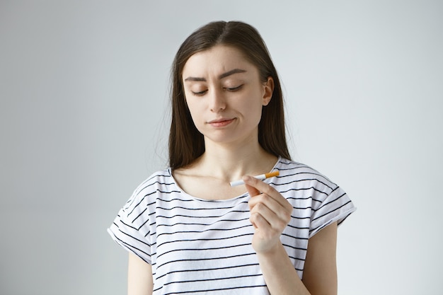 stirnrunzelnde junge Frau in gestreiftem Oberteil hält unbeleuchtete Zigarette in der Hand und betrachtet sie mit enttäuschtem Ausdruck